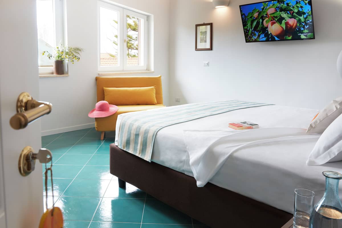 A bedroom with a bed, a bedside table with a glass and a bottle, a window with a view of trees, a chair with a hat, and a picture on the wall.