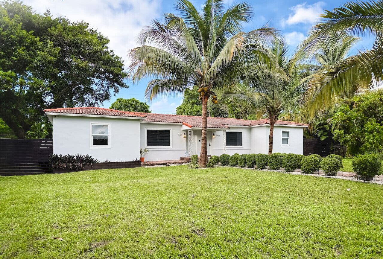 A white house with a red-tiled roof, a well-maintained lawn, and several palm trees.