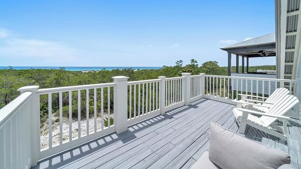 A wooden deck with white railings, two Adirondack chairs, and a view of the ocean and greenery.