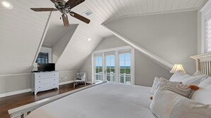 A bedroom with a large bed, a ceiling fan, a dresser, a chair, and a balcony with a view.