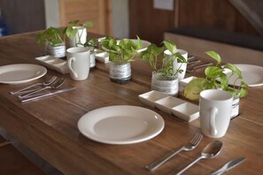 Une table de salle à manger en bois, mise avec de l’assiette blanche, des couverts et des tasses blanches, chaque place étant agrémentée d’une petite plante en pot.