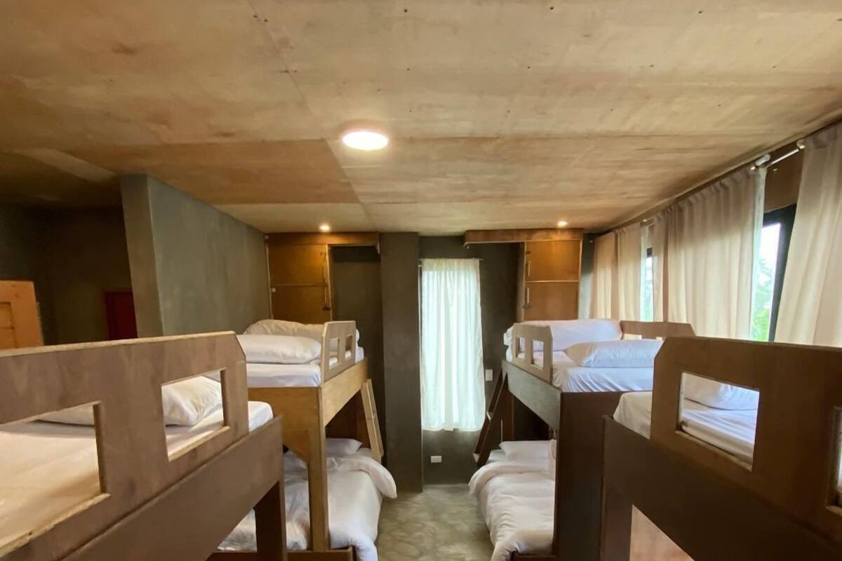 A bunk room with wooden bunk beds, a central walkway, and a window with curtains.