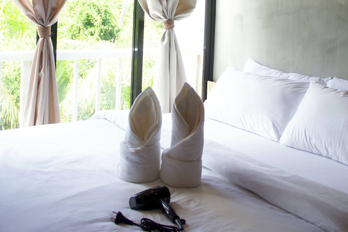A hotel room with a bed, white curtains, and a window with a view of greenery.