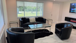 A modern living room with a sofa, two armchairs, a glass coffee table, and a TV mounted on the wall. - Charming 2-bedroom house in Charlotte Harbor, walk to restaurants & shopping (Port Charlotte)