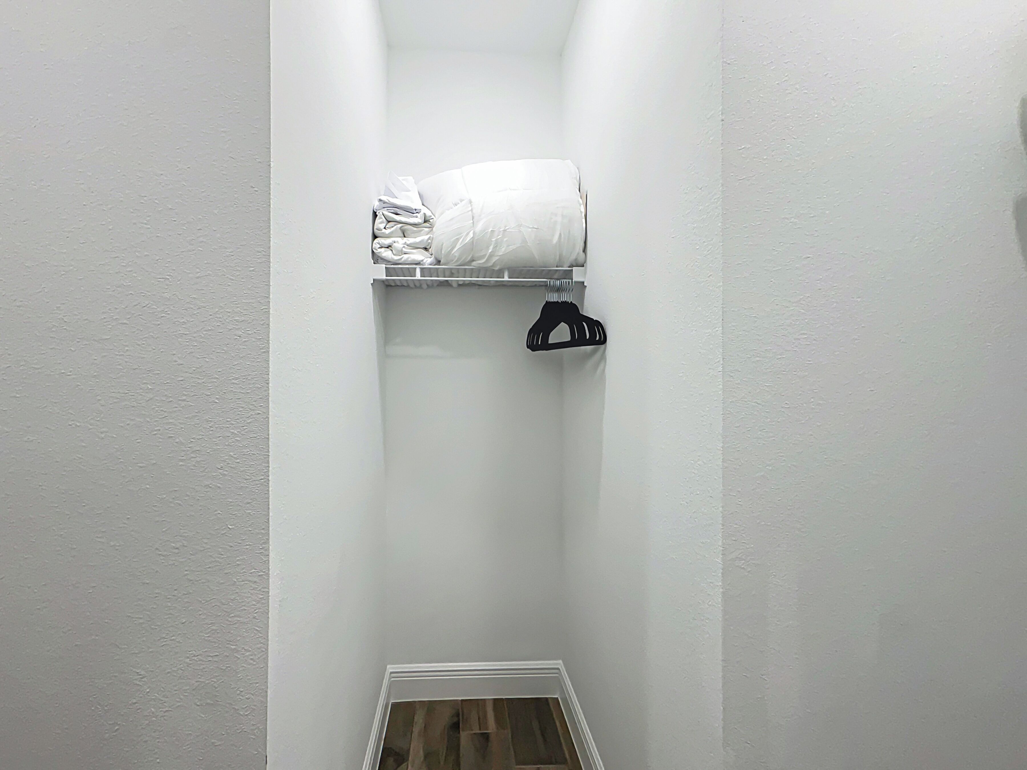 Una habitación pequeña y estrecha con pared, puerta y techo blancos. Se observa una repisa blanca con una manta blanca doblada y un perchero negro.