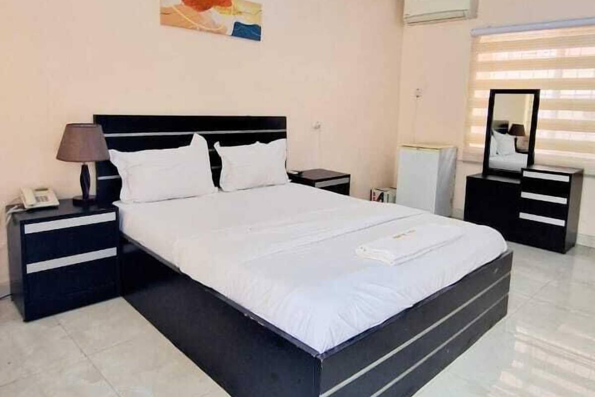 A modern hotel room with a large bed, a nightstand with a lamp, a dresser, and a mirror.