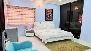 A modern bedroom with a bed, a blue armchair, a nightstand, a wall-mounted TV, and a large window with blinds. - Habitat Suites and Apartments (Lagos)