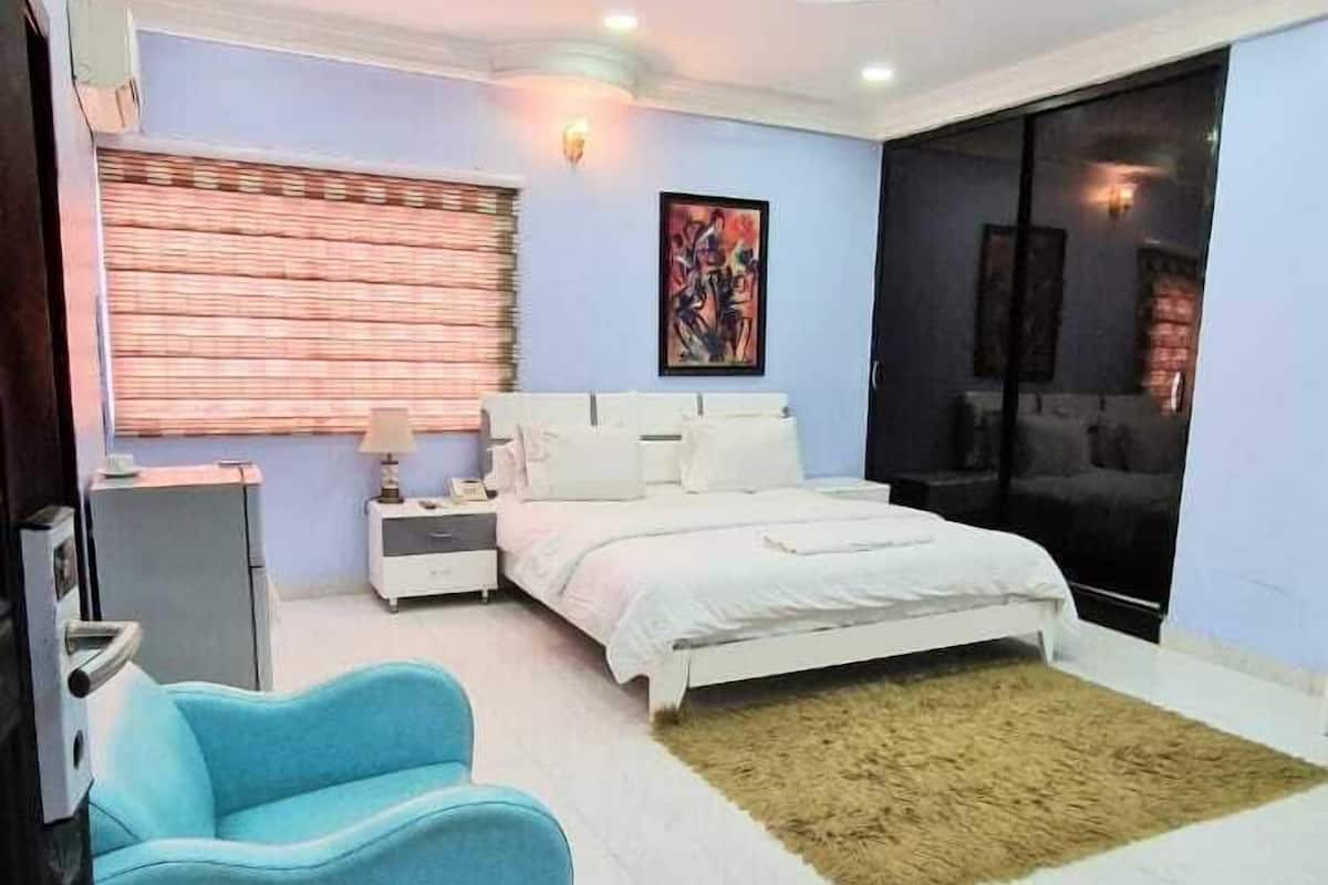 A modern bedroom with a bed, a blue armchair, a nightstand, a wall-mounted TV, and a large window with blinds.
