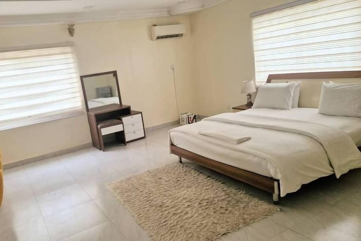 A bedroom with a bed, a bedside table, a mirror, a dresser, and a window with blinds.