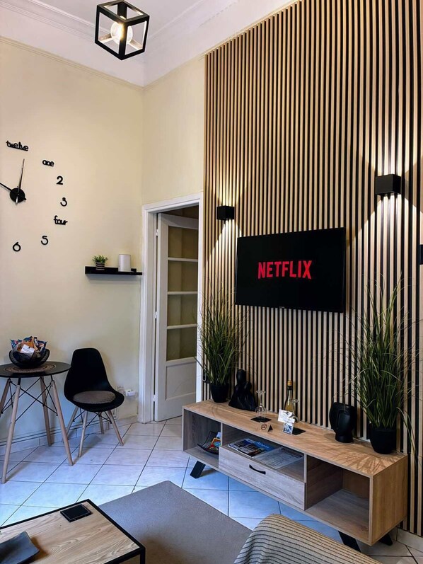 Una sala de estar moderna con un muro de listones de madera, un televisor montado que muestra 'NETFLIX', un reloj de pared y una pequeña mesa con una cesta.