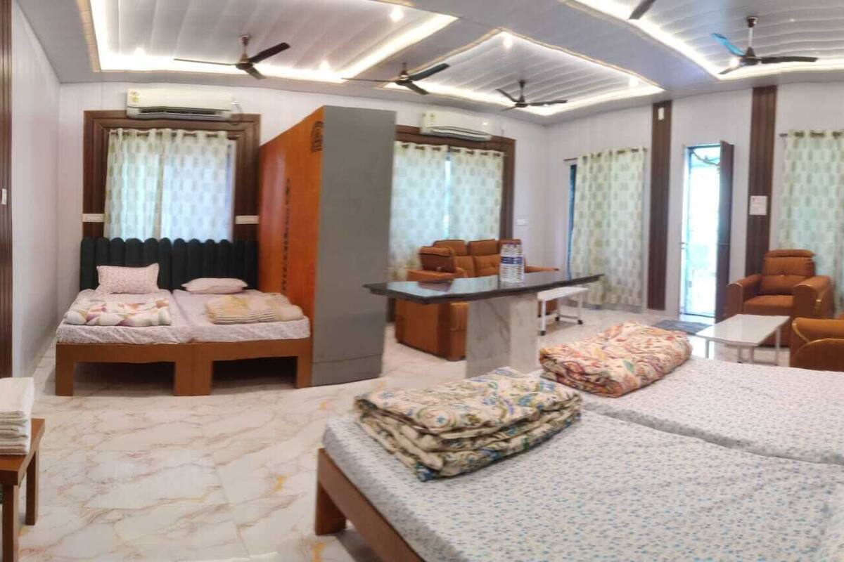 A hotel room with a bed, a sofa, a desk, and a chair. There are curtains on the windows and an air conditioning unit on the wall.