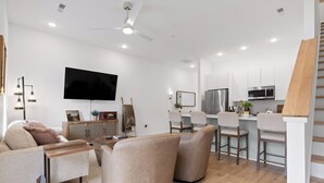 Una sala de estar moderna con sofá, sillón y una mesa de centro. Hay un televisor de pantalla plana montado en la pared, un ventilador de techo e iluminación empotrada.