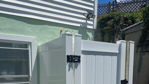 Una estructura exterior blanca con una puerta y una ventana, posiblemente un cobertizo o cuarto de servicio, junto a una casa con revestimiento verde y una ventana blanca.