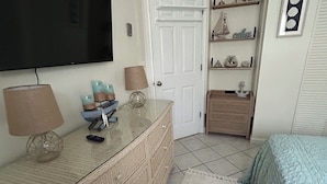 Un dormitorio con televisión, cómoda, cama y estante con objetos decorativos.