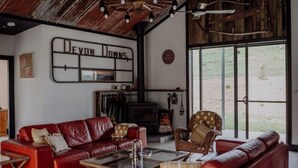 Amplia sala de estar con un sofá de cuero rojo, una silla de mimbre y una mesa de centro de vidrio. Se observa una chimenea con repisa, y la habitación tiene un techo rústico con vigas a la vista.