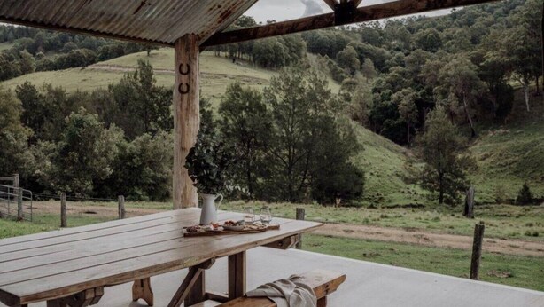 Una mesa de madera rústica con una jarra blanca y un plato de comida, frente a un paisaje montañoso con árboles.