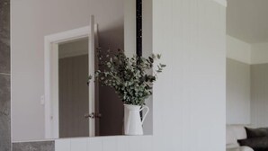 Canto de um quarto com uma cesta de vime contendo lenha, um vaso branco com folhagem verde e uma porta branca.