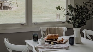 Uma mesa de jantar com um pão, uma xícara, um vaso com plantas e uma janela com vista para uma área gramada.