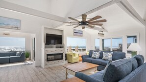 A spacious living room with a large window offering a view of the city, a ceiling fan, and a fireplace. - Oceanfront charm, heated pool, hot tub, dog friendly, 4 bed 3.5 bath (Frisco)