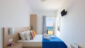 A hotel room with a bed, a bedside table with a lamp, a TV mounted on the wall, and a view of the beach. - Creta Beachfront Apartment B for 2 persons by MPS (Paralia Fodele)