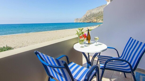 A balcony with two striped chairs, a small round table with a bottle and glasses, and a view of the beach and sea. - Creta Beachfront Apartment B for 2 persons by MPS (Paralia Fodele)