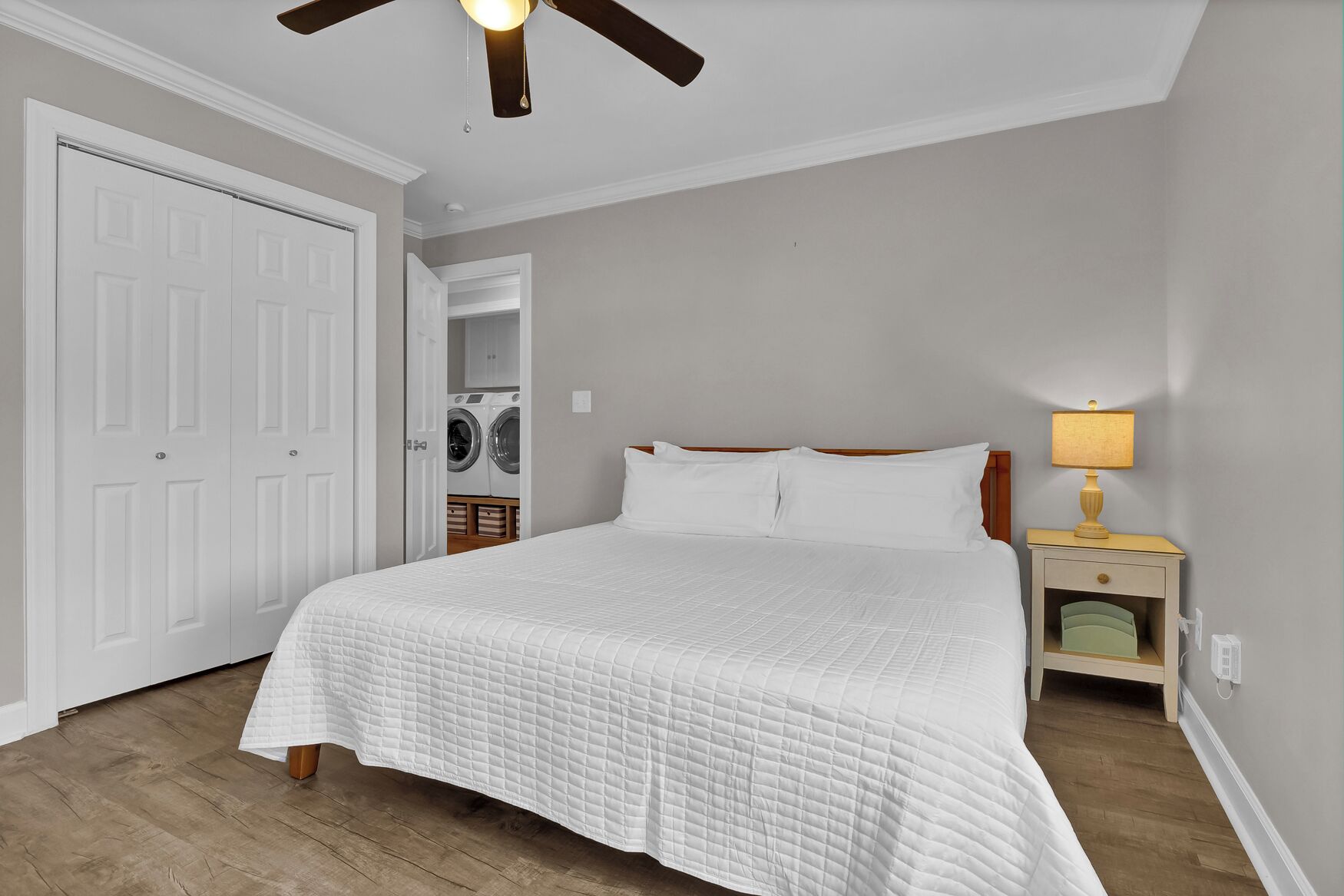 A bedroom with a bed, a nightstand, a lamp, a ceiling fan, and a laundry room visible through an open door.
