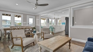 Un vaste salon comprenant un grand canapé, deux fauteuils, un coin repas avec une table et des chaises en bois, ainsi qu’une cuisine dotée de meubles blancs et d’un grand îlot central.