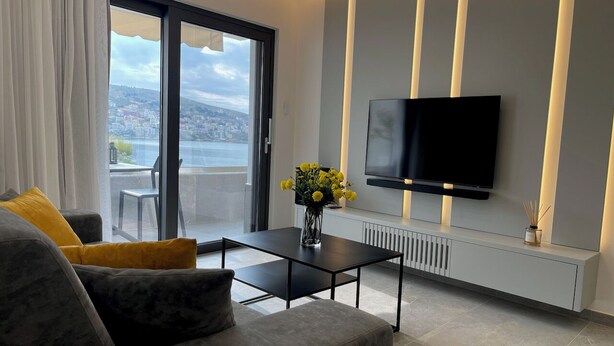 A modern living room with a sofa, a coffee table, a flat-screen TV, and a view of a cityscape through a large window. - Italian design apartment for a refined explorer of new destinations. (Saranda)