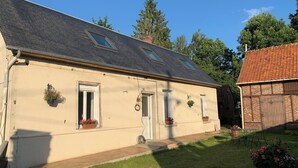 Une maison de plain-pied avec un toit en pente, une porte d’entrée et deux fenêtres. Des plantes en pot sont posées sur le rebord de la fenêtre, et un petit jardin avec de l’herbe et une clôture en bois se trouve à l’avant.