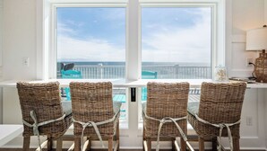 A room with a view of the ocean through large windows, wicker chairs, and a white table.