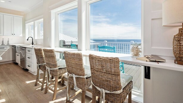 A modern kitchen with white cabinetry, a large window overlooking the sea, and wicker bar stools.