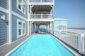 A poolside area with a view of the beach, a covered deck, and a two-story building with a balcony. - Seaside Delight: Luxury Oceanfront Home with Pool, Elevator (Oak Island)