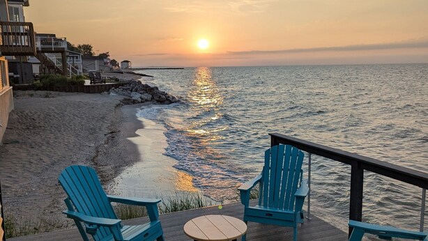 A wooden deck with four blue Adirondack chairs and a small round table, overlooking a beach and the ocean at sunset.