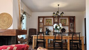 Una sala de estar acogedora con una mesa de comedor de madera, sillas y una chimenea. Hay objetos decorativos en la repisa de la chimenea y una araña colgante sobre la mesa.