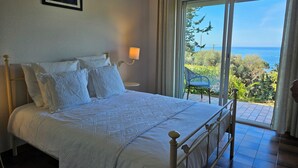Un dormitorio con cama, almohadas, una mesita de noche, una lámpara y vistas al mar a través de un ventanal grande.