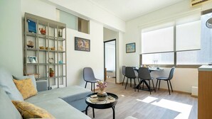 A modern living room with a sofa, a dining area with chairs, a bookshelf, and a window providing natural light. - Athenais 3A 2 bedroom 4 persons apartment by MPS (Athina)