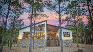 A modern cabin with a stone foundation, large windows, and a wooden deck. - 6BR Luxe Cabin w/ Pool & Pickleball | Broken Bow (Broken Bow)