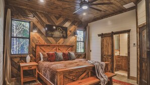 Um quarto rústico com teto de madeira, uma cama grande com várias almofadas, uma mesa de cabeceira, um banco e um espelho grande.