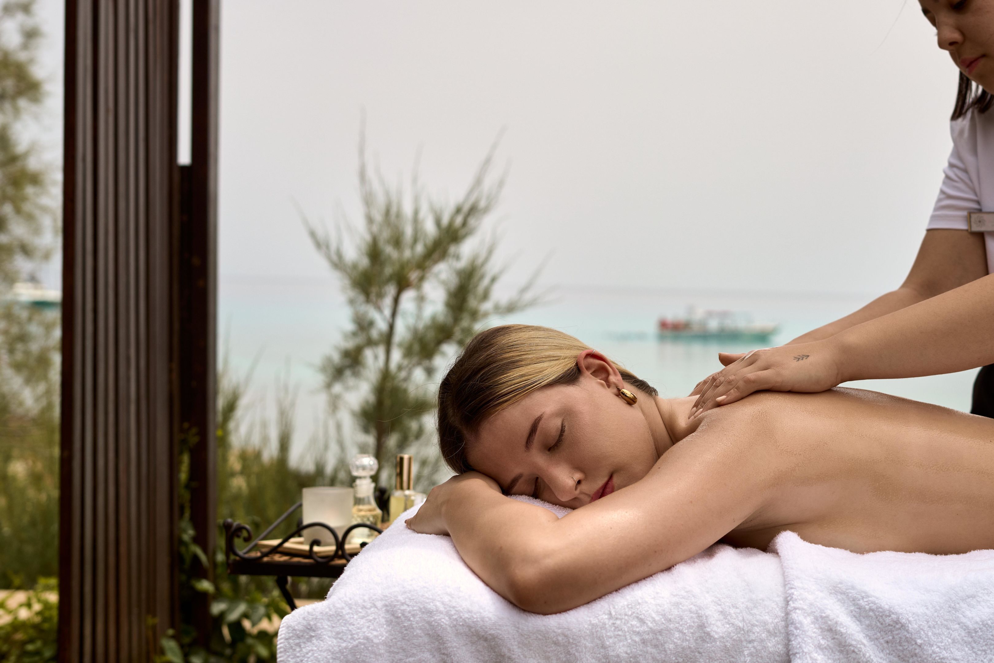 A person receiving a back massage outdoors with a view of the sea and greenery in the background.