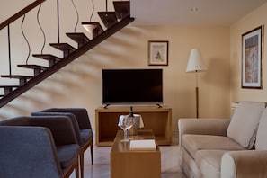 A modern living room with a wooden staircase, a flat-screen TV, a sofa, and a coffee table with a bottle and glass. - Lindian Dream Seaside Resort (Lindos)