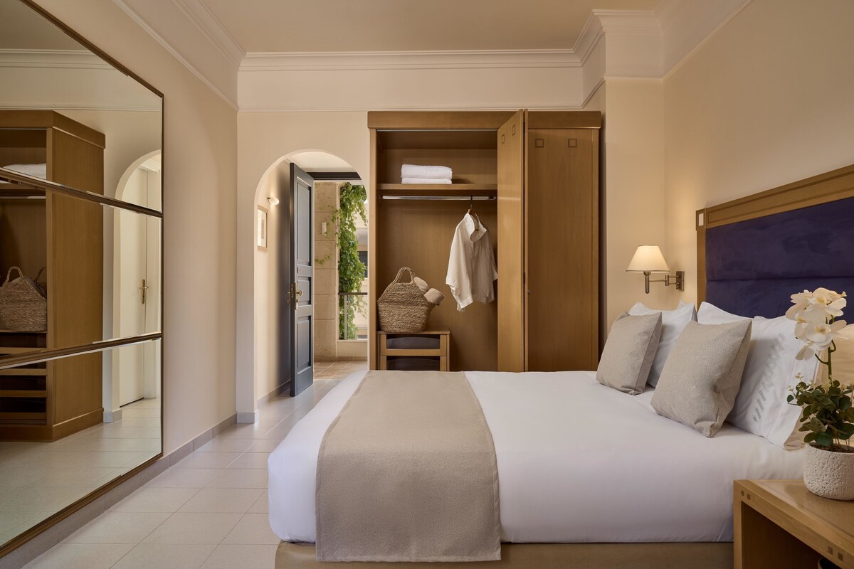 Habitación de hotel con una cama grande, un armario, un espejo y una mesita de noche con una lámpara y una planta.
