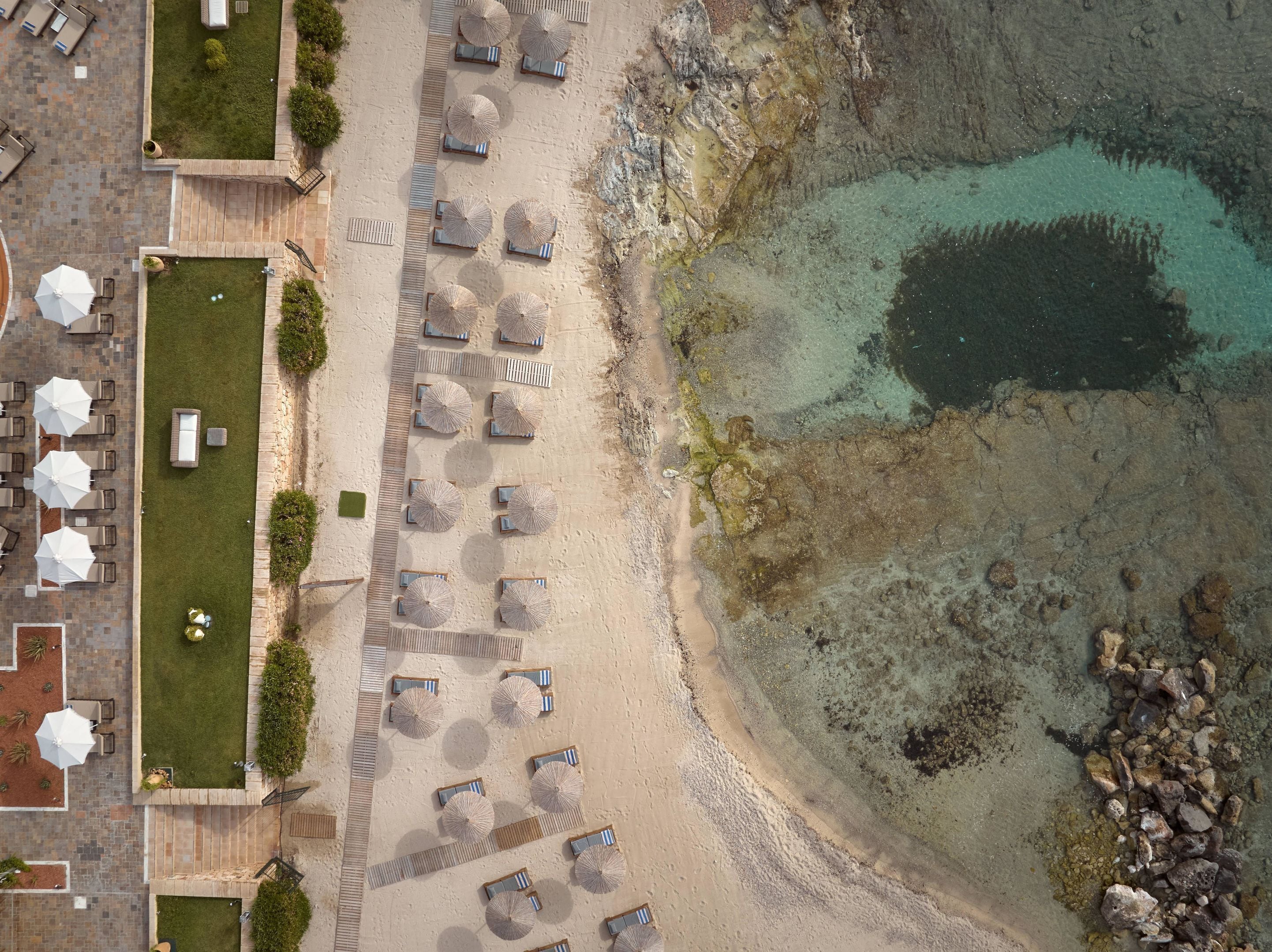 Vue aérienne d’un complexe hôtelier en bord de mer, avec des rangées de chaises longues et de parasols, un espace piscine et une côte rocheuse.
