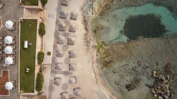 Vue aérienne d’un complexe hôtelier en bord de mer, avec des rangées de chaises longues et de parasols, un espace piscine et une côte rocheuse.