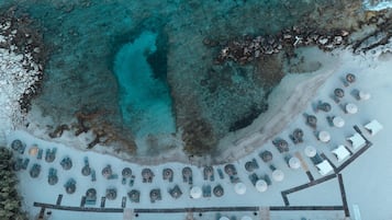 Vue aérienne d’un complexe hôtelier en bord de mer, avec une eau turquoise cristalline, une plage de sable et des rangées de transats et de parasols.