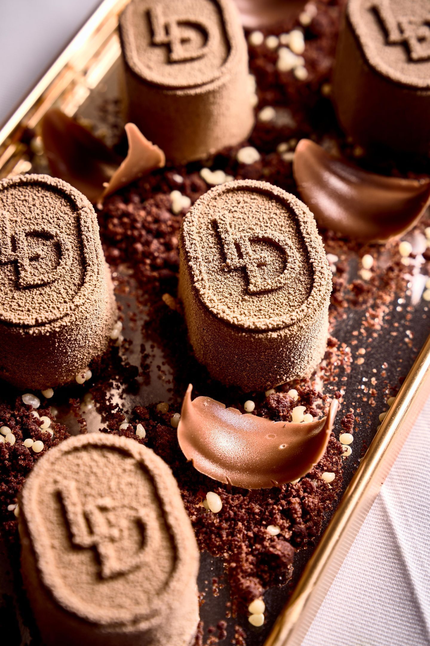 Una bandeja de postres bañados en chocolate con un logotipo 'D', sobre una superficie oscura y migas dispersas.