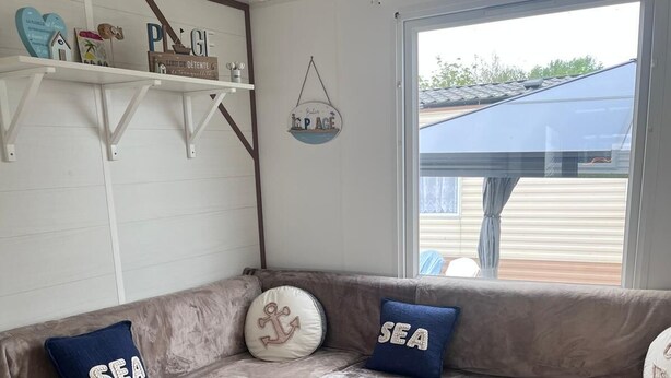 A cozy living area with a beige sectional sofa, two pillows with 'SEA' text, a small white coffee table, and a window with a view of a house.