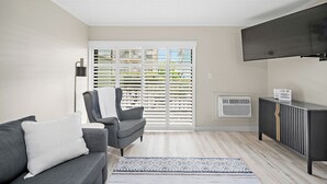 Un salon avec un canapé gris, un fauteuil, un téléviseur à écran plat, un meuble et une fenêtre avec des stores.