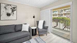 Un salon moderne avec un canapé gris, un tapis à motifs, un fauteuil sur lequel est posée une serviette blanche, un lampadaire noir et un grand tableau encadré représentant un cheval.