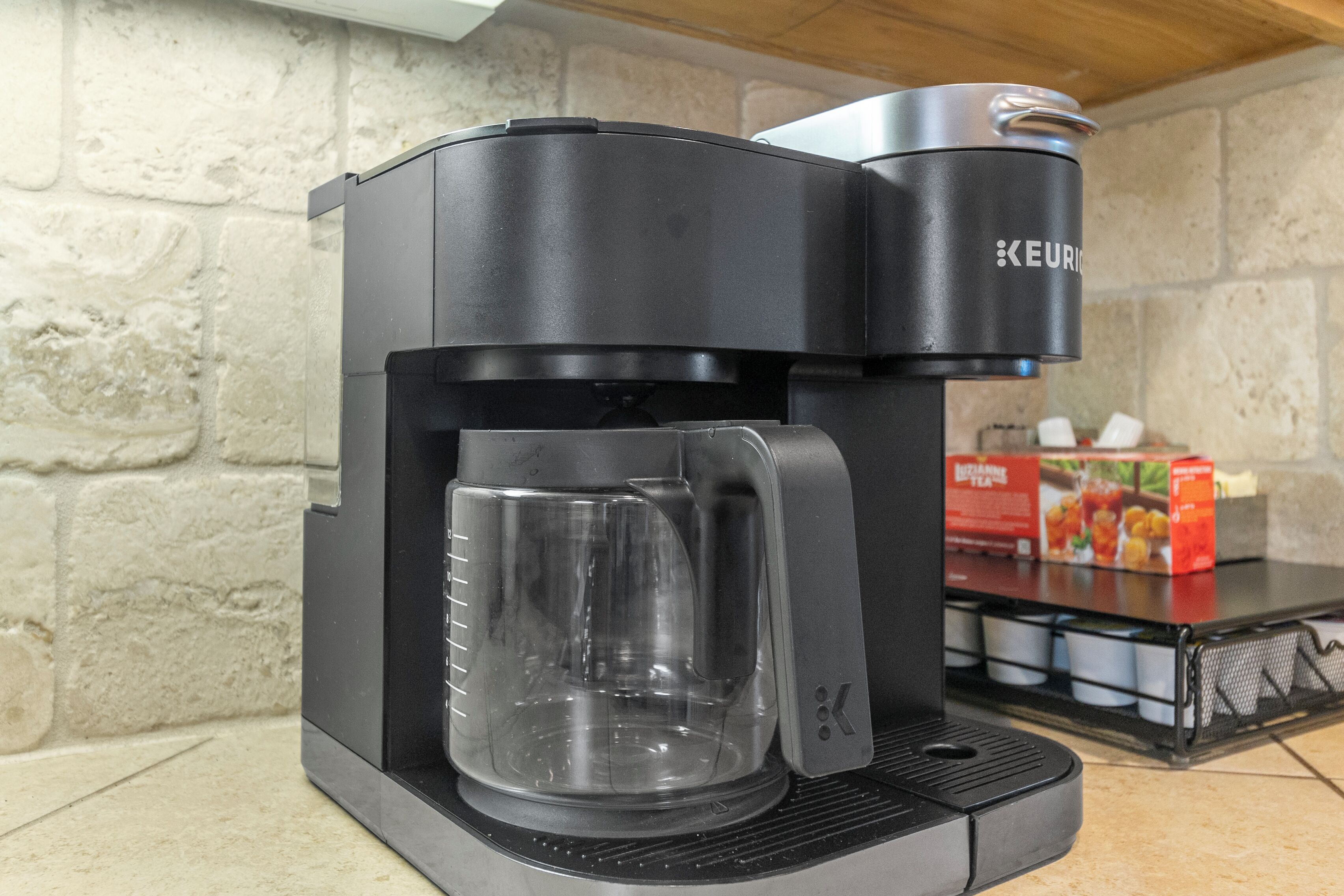 A Keurig coffee maker with a clear mug on its base, a stone wall background, and a box of coffee pods on a shelf.