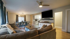 A modern living room with a large sectional sofa, a dining area with a table and chairs, a kitchen with white cabinets, and a ceiling fan.